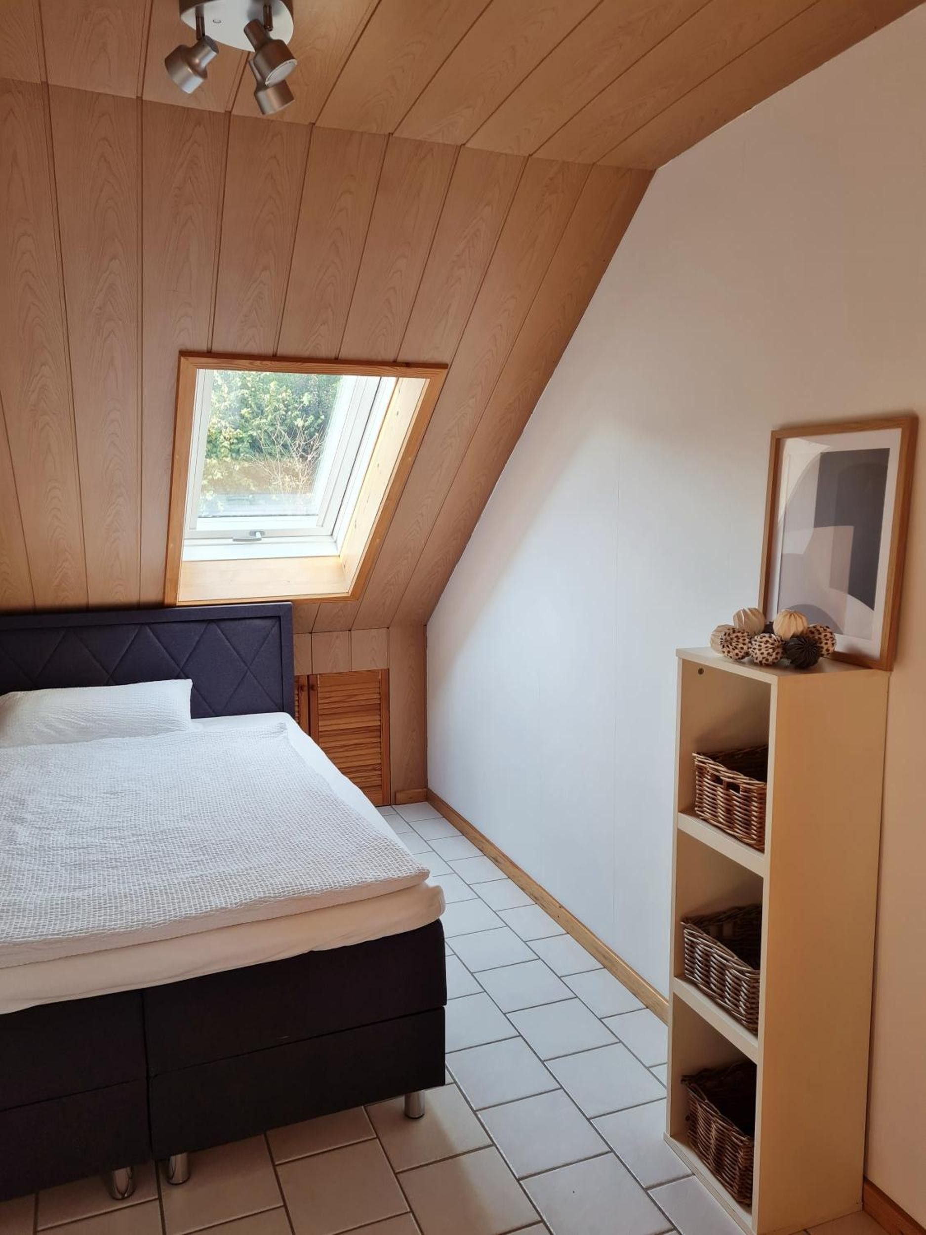 Een slaapkamer met een schuine kap, dakraam, een bed, een nachtkastje en een wandplank met manden.
