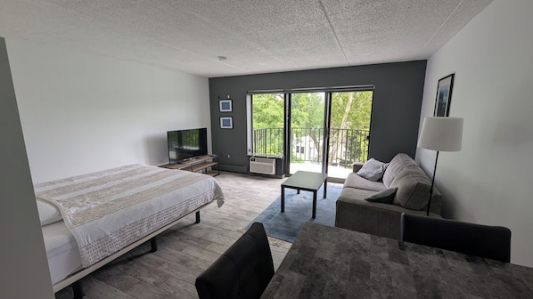 Habitación de hotel moderna con cama, sofá, televisión y balcón con vistas a árboles.