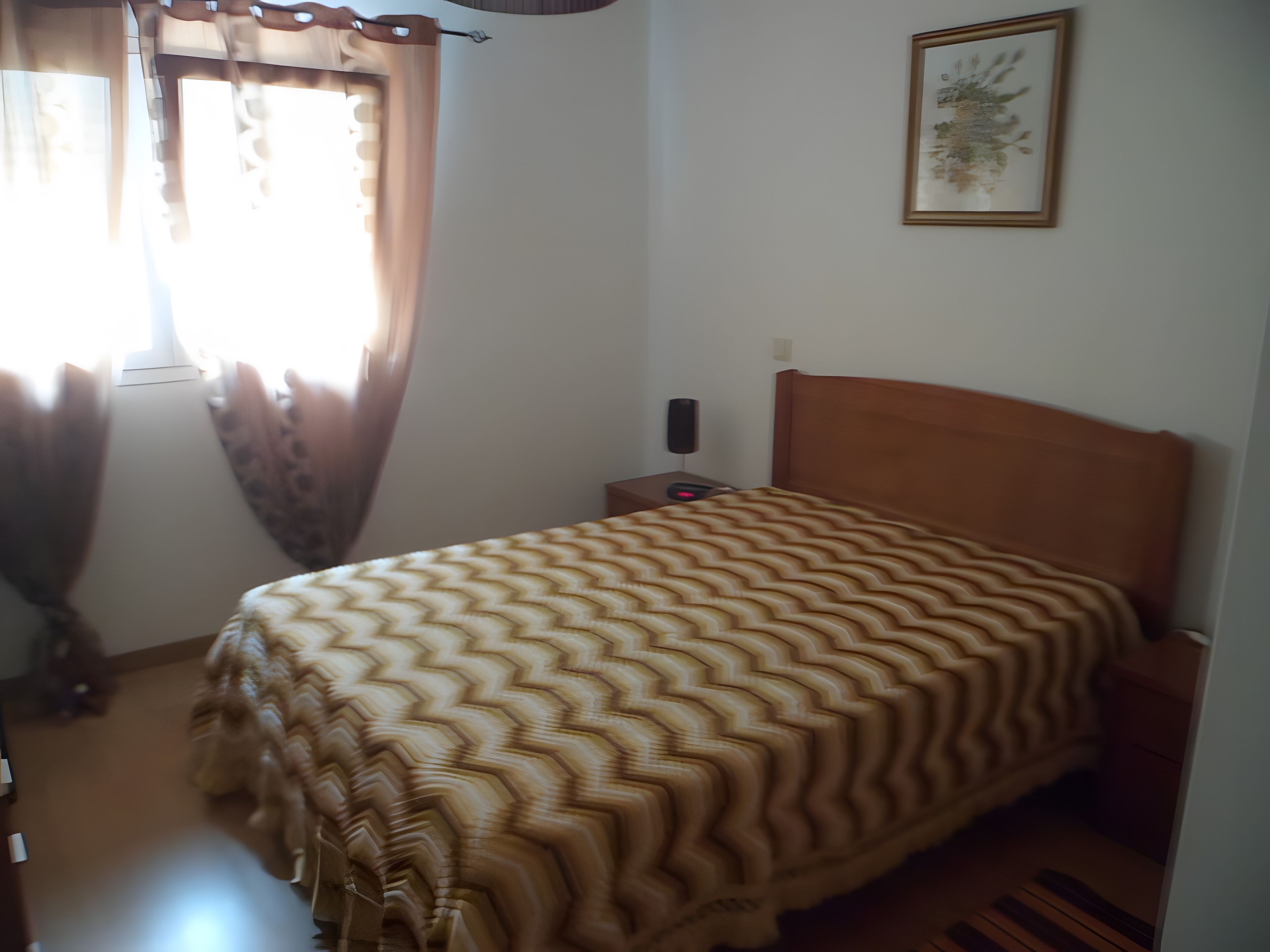 Een slaapkamer met een bed, gordijnen, een nachtkastje en een aan de muur hangend schilderij.