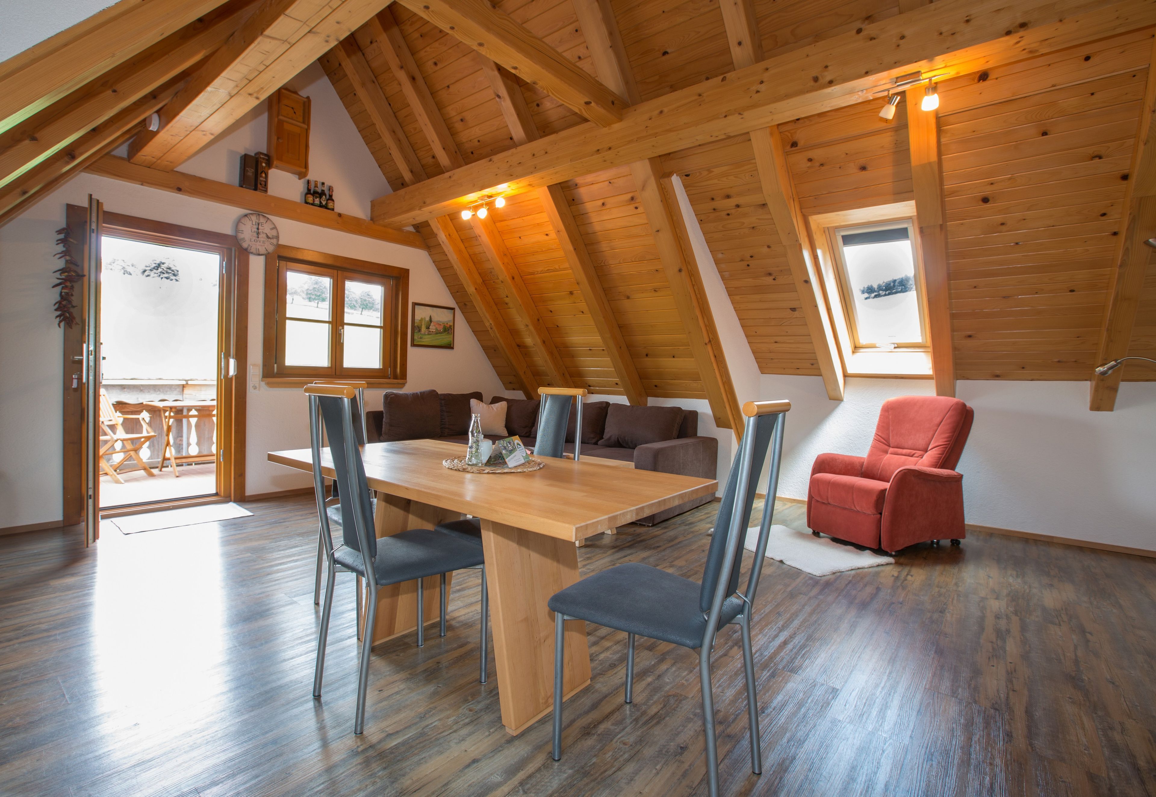 Un coin repas avec une table et des chaises en bois, un fauteuil rouge et une vue sur l’extérieur par une fenêtre.