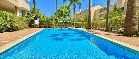 Ein klares Schwimmbecken, umgeben von Palmen und einem gepflasterten Weg, mit Gebäuden im Hintergrund.