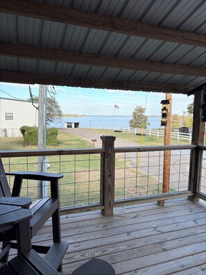 A wooden deck with a metal railing overlooking a body of water, a flag, and a building in the background. - Eagle Lake Hunting and Fishing Cabin (Vicksburg)