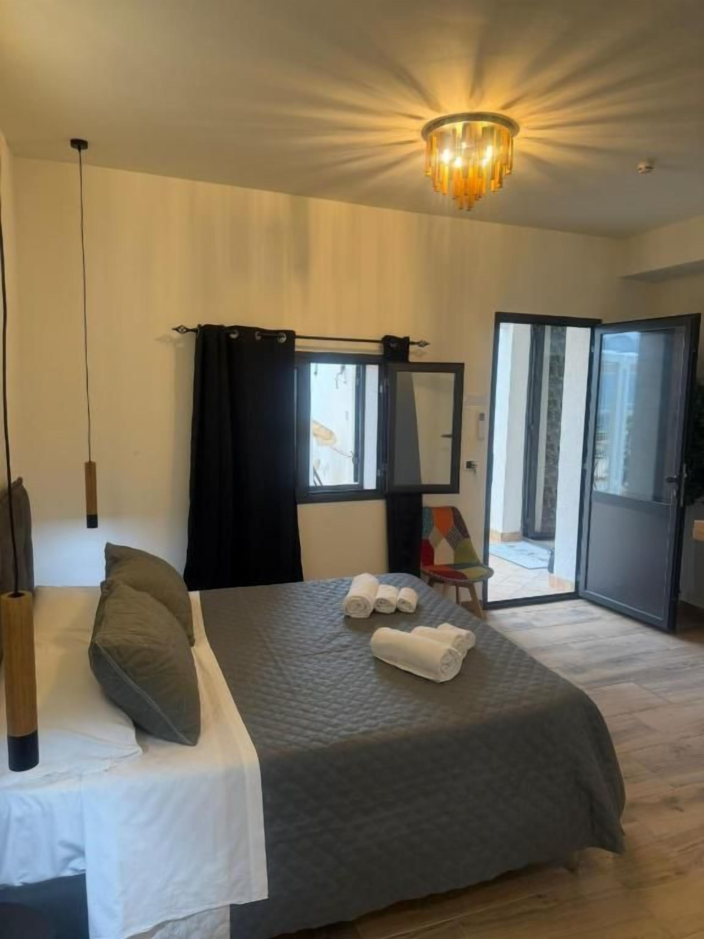 A hotel room with a bed, a large mirror, a chandelier, and a view through an open door.