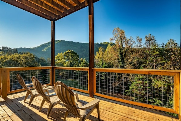 Um deck de madeira com duas cadeiras e uma mesa, com vista para uma paisagem arborizada com colinas.