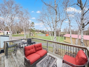 A deck with outdoor furniture, including a red sofa and chairs, overlooking a grassy area with trees and a building in the background. - Unit A/ Spacious/ Heated pool/ Close to beach/ On-site Activities! (Brackley Beach)