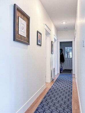 A hallway with framed artwork on the walls, a patterned rug, and a door leading to another room. - Unit A/ Spacious/ Heated pool/ Close to beach/ On-site Activities! (Brackley Beach)