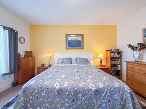 A bedroom with a bed, two bedside tables, a nightstand, a bookshelf, a clock on the wall, and a window with curtains. - Unit A/ Spacious/ Heated pool/ Close to beach/ On-site Activities! (Brackley Beach)