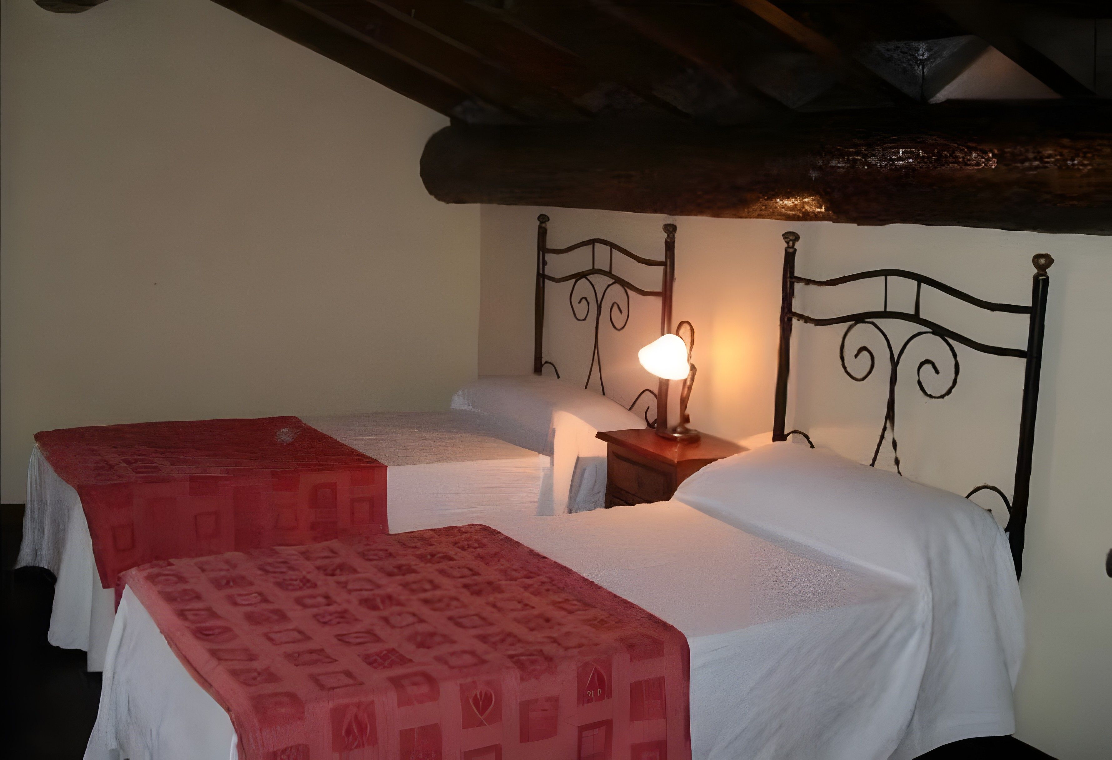Une chambre avec deux lits simples, une tête de lit en bois, une table de chevet avec une lampe et une couvre-lit rouge à motifs.