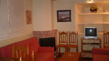 Un salon avec un canapé rouge, des chaises en bois, une cheminée, un téléviseur posé sur un meuble en bois et un tableau encadré accroché au mur.