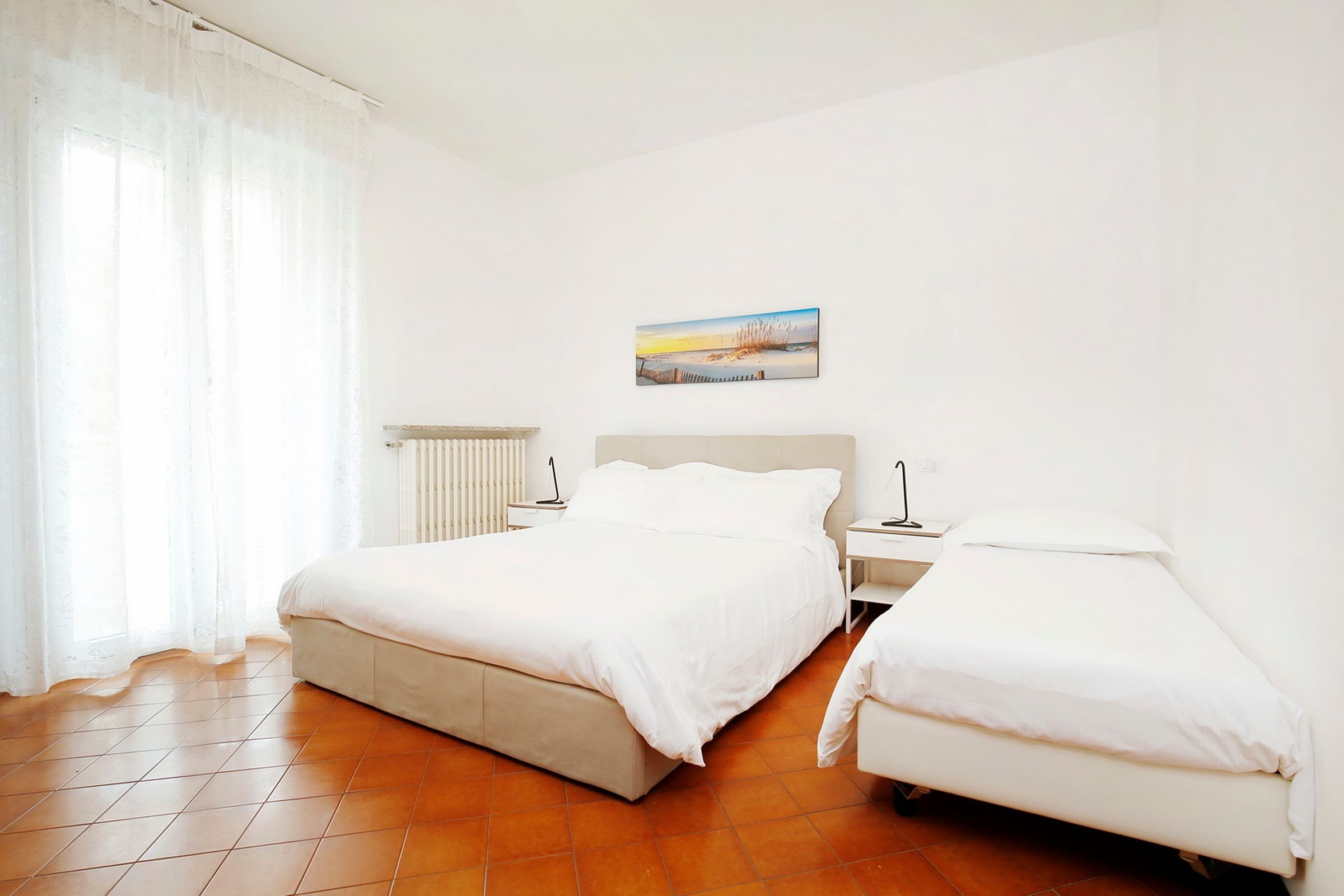 Una habitación de hotel con una cama, una cama individual, una mesita de noche, una lámpara, un radiador y un cuadro en la pared.