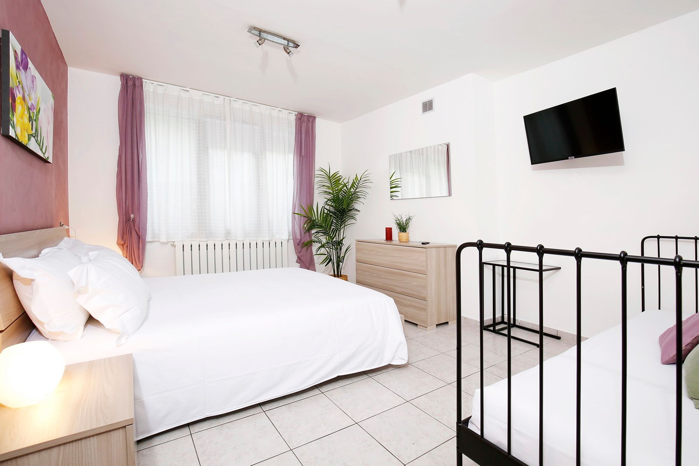 Un dormitorio moderno con cama, mesita de noche, cómoda, planta en maceta y televisor montado en la pared.