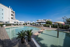Piscina con sillones de descanso y sombrillas, rodeada por un edificio moderno con balcones.