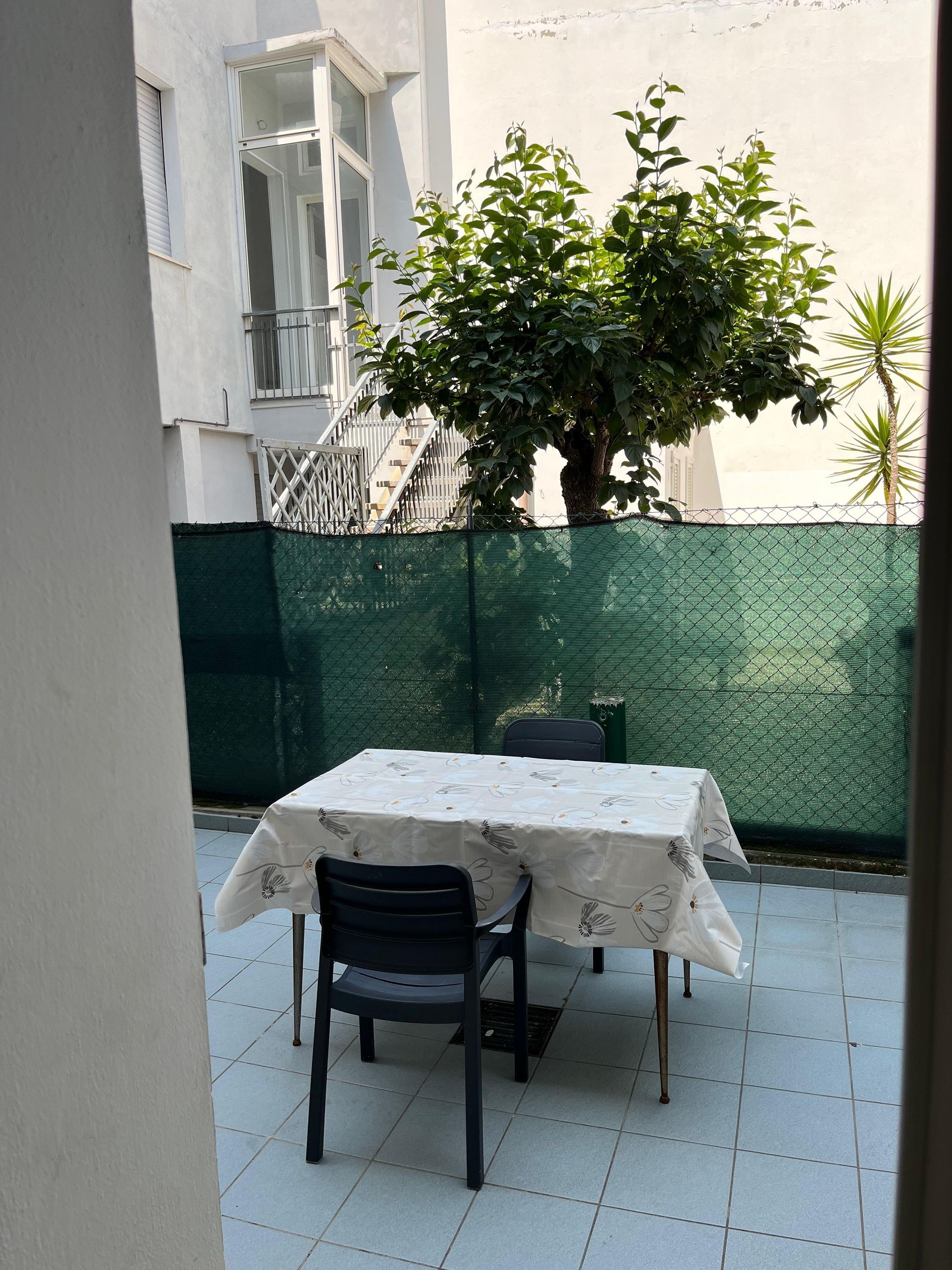 A patio with a table and chairs, a green fence, and a tree.