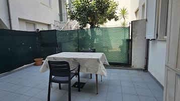 Pequeno alpendre ao ar livre com mesa e cadeiras, cercado por um muro verde e um edifício branco.