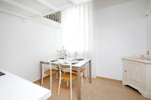 Un comedor con una mesa blanca para cuatro personas, un florero con flores y un armario blanco con velas.