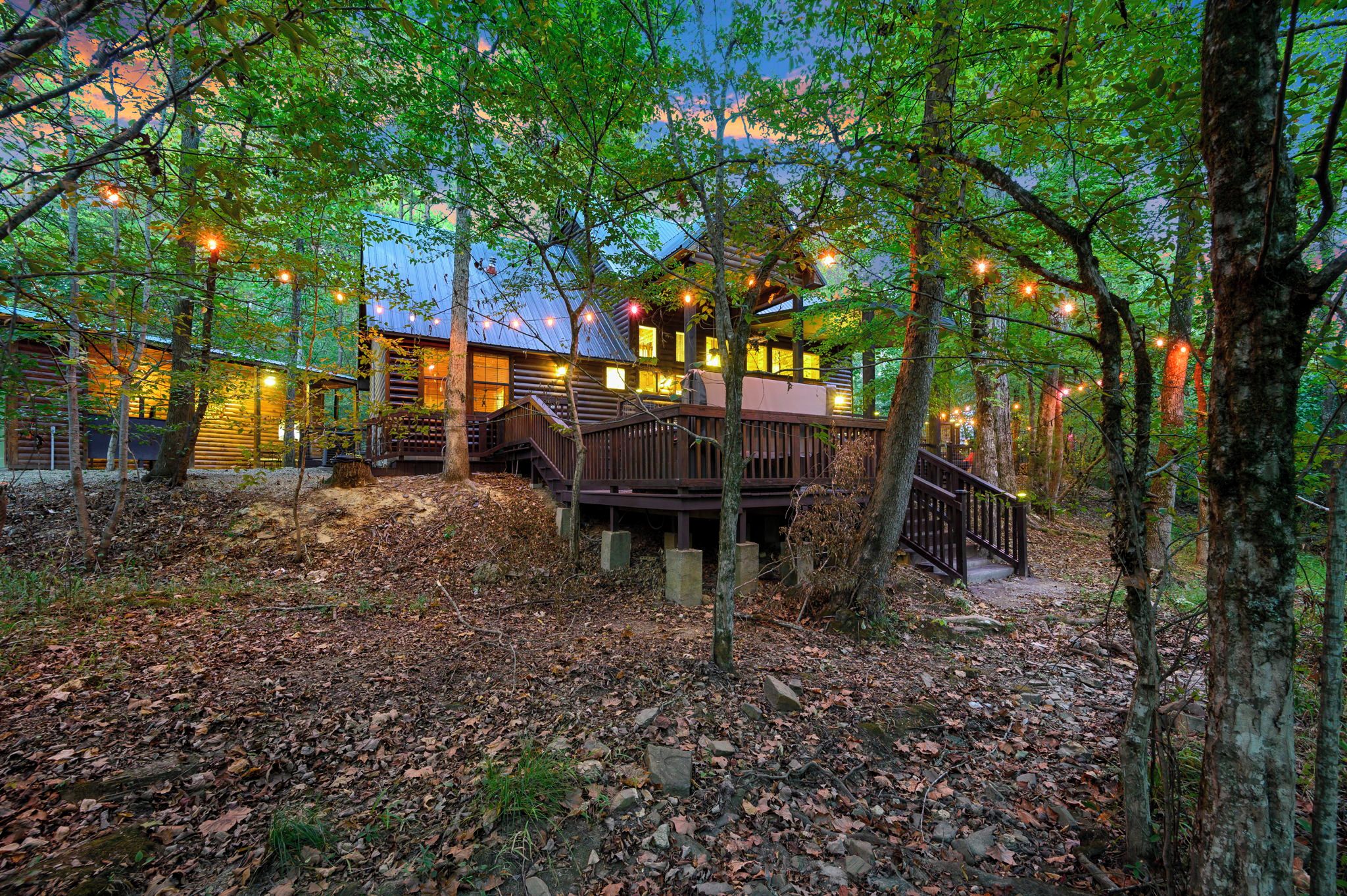 A cabin with a wooden deck, surrounded by trees and a forest floor covered with leaves.