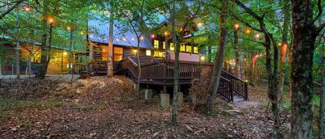 A cabin with a wooden deck, surrounded by trees and a forest floor covered with leaves.