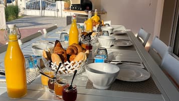 An outdoor dining area with a long table set for a meal, including bread, condiments, and beverages.
