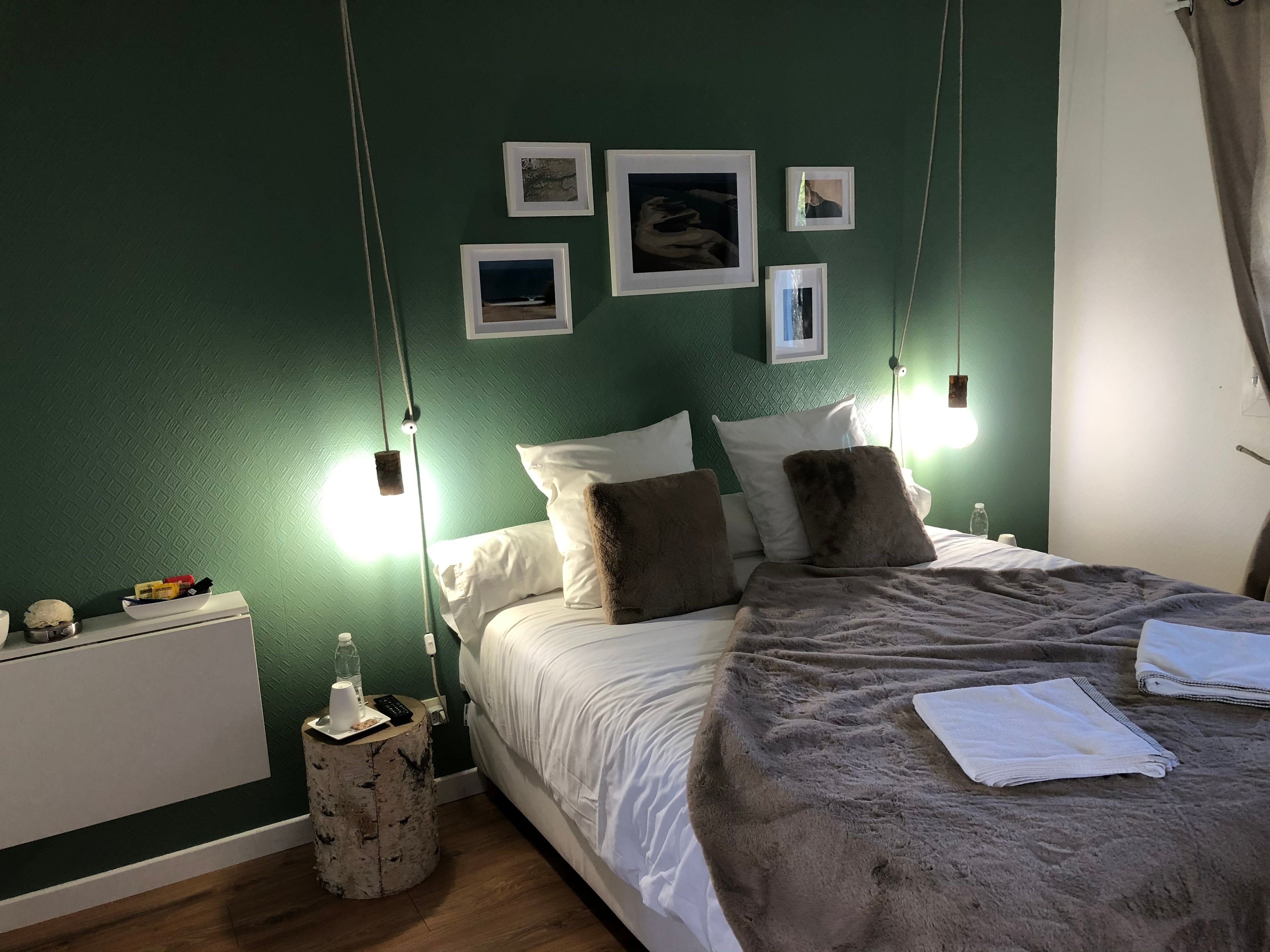 A neatly made bed with pillows, a bedside table with a bottle and a remote, and a wall with framed pictures.