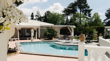 A swimming pool with a covered patio area, outdoor seating, and a view of trees.