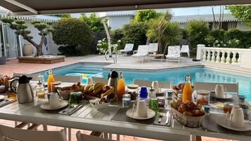 A poolside breakfast setup with a table, chairs, and a view of the pool and garden.