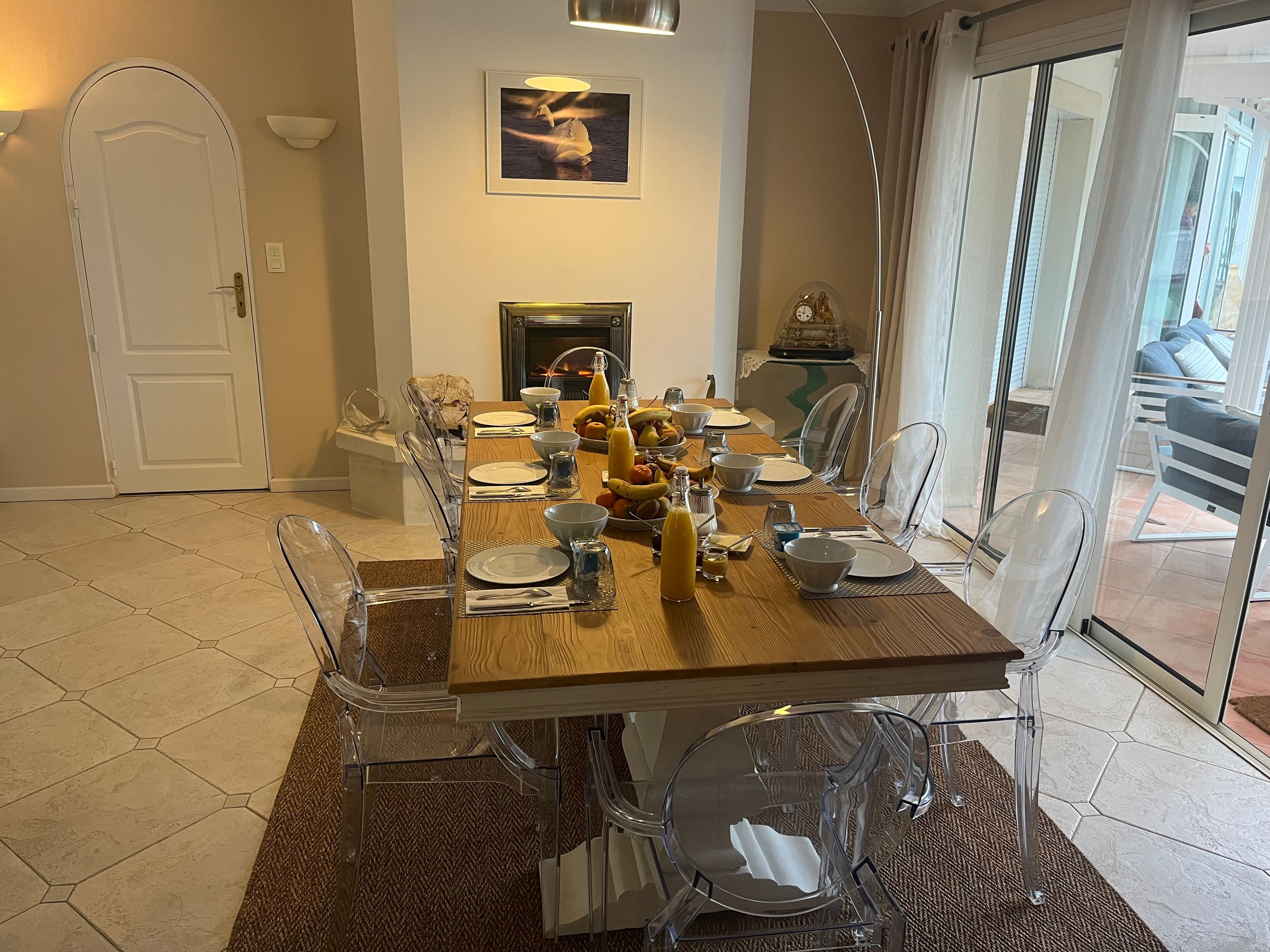 A dining area with a wooden table set for four, surrounded by clear chairs, a fireplace, and a view through a glass door.