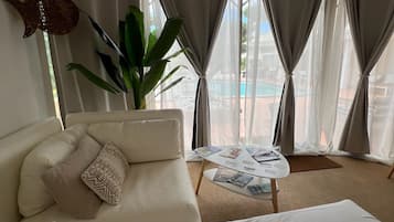 A hotel room with a bed, a sofa, a small table with magazines, a plant, and a view of a pool.