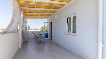 A covered outdoor balcony with a table and chairs, a window, and a door leading to an interior space.