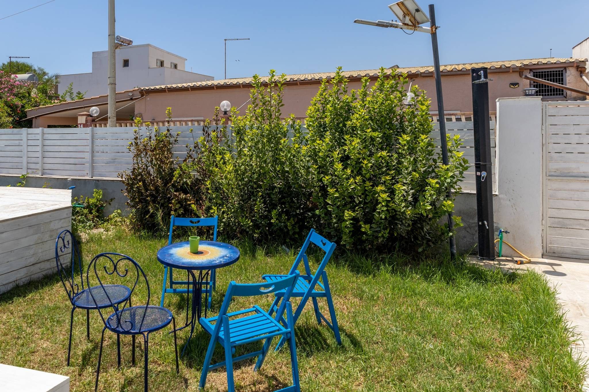 Zona exterior amb mobiliari per seure, taula i cadires, envoltada de vegetació i una tanca.
