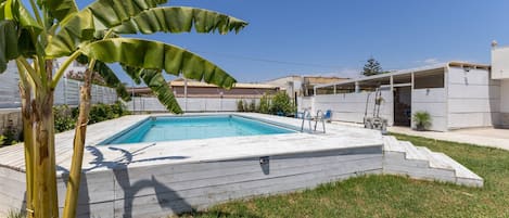 Une piscine avec une terrasse en bois, une échelle et un palmier au premier plan.