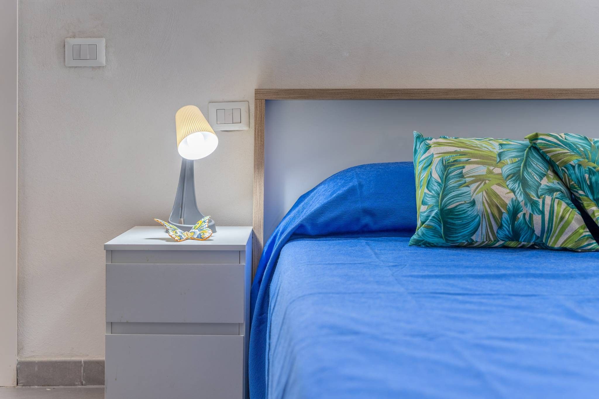 A bedroom with a bed, a nightstand with a lamp, and a wall switch.