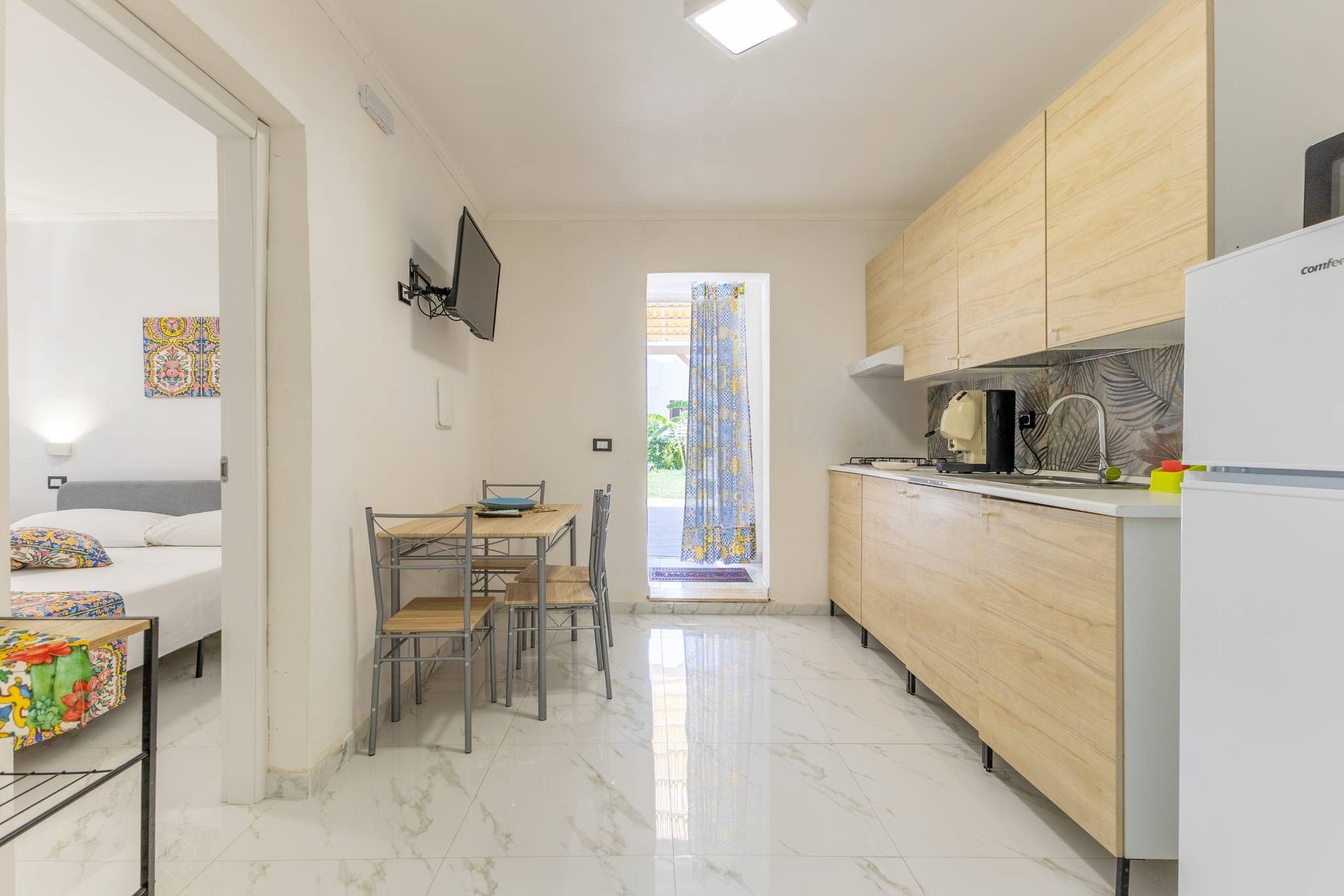 A compact hotel room with a bed, a desk, chairs, a television, and a kitchenette.