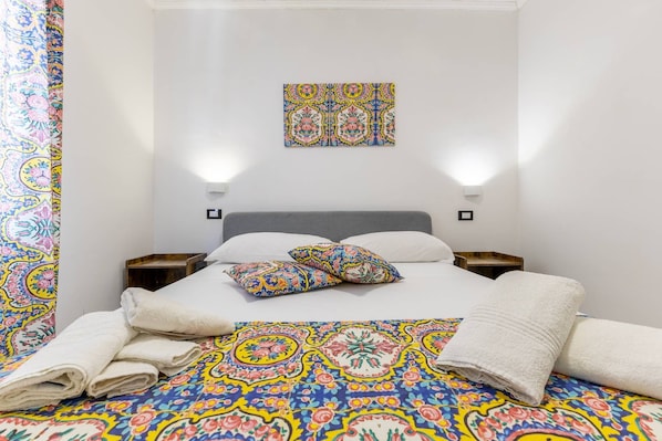 Ett hotellrum med en säng, kuddar, ett sängbord, ett konstverk som hänger på väggen och ett mönstrat sängöverkast.