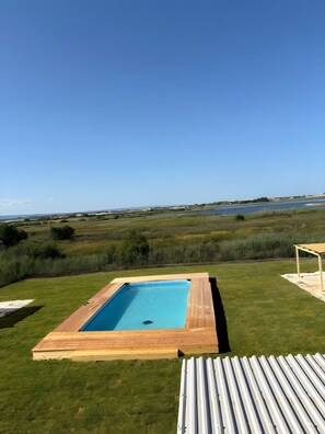 Piscina rectangular con plataforma de madera, rodeada de césped y vista a un cuerpo de agua y terreno distante.