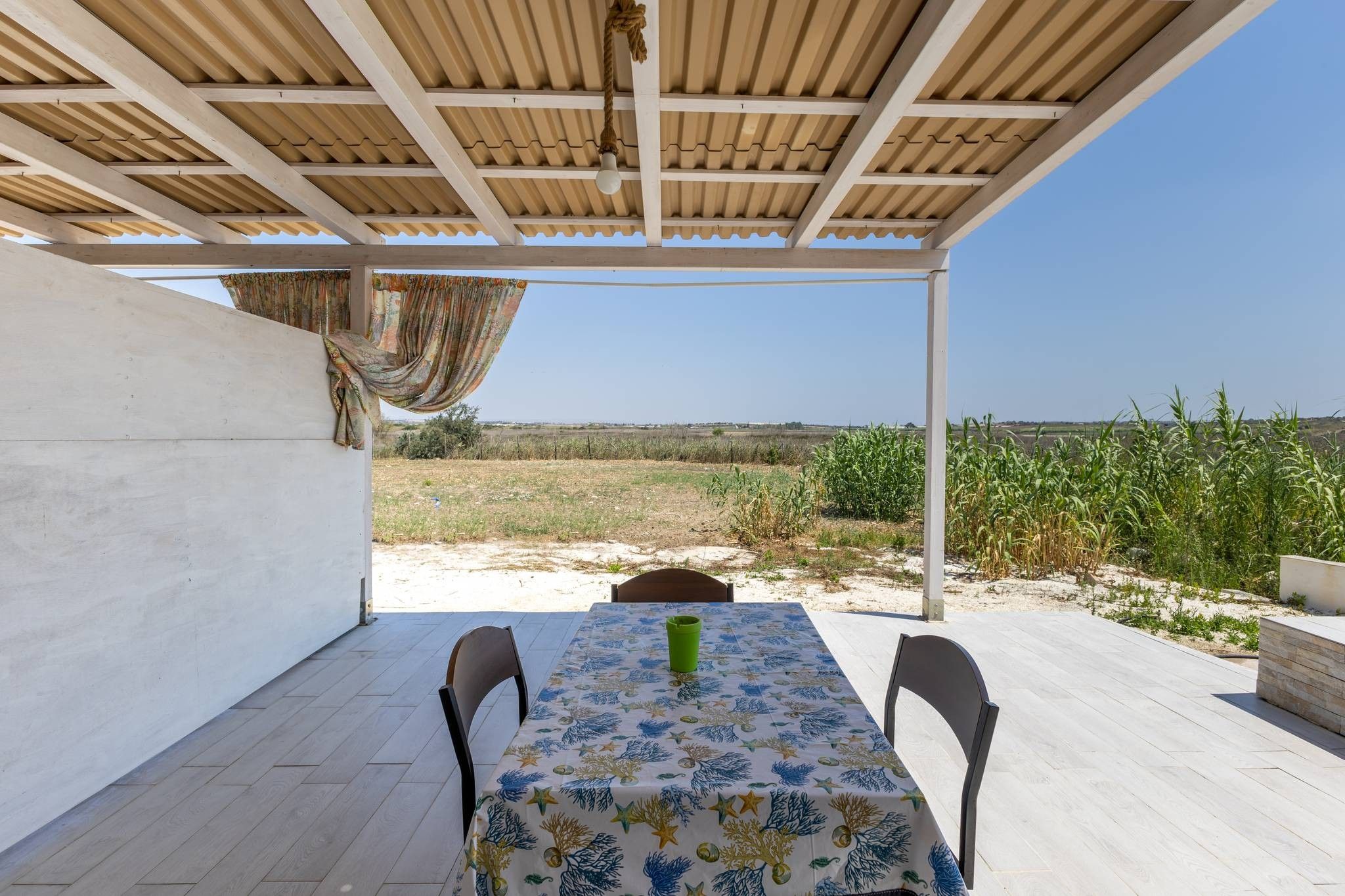 A covered outdoor dining area with a table and chairs, a view of a field, and a clear sky.