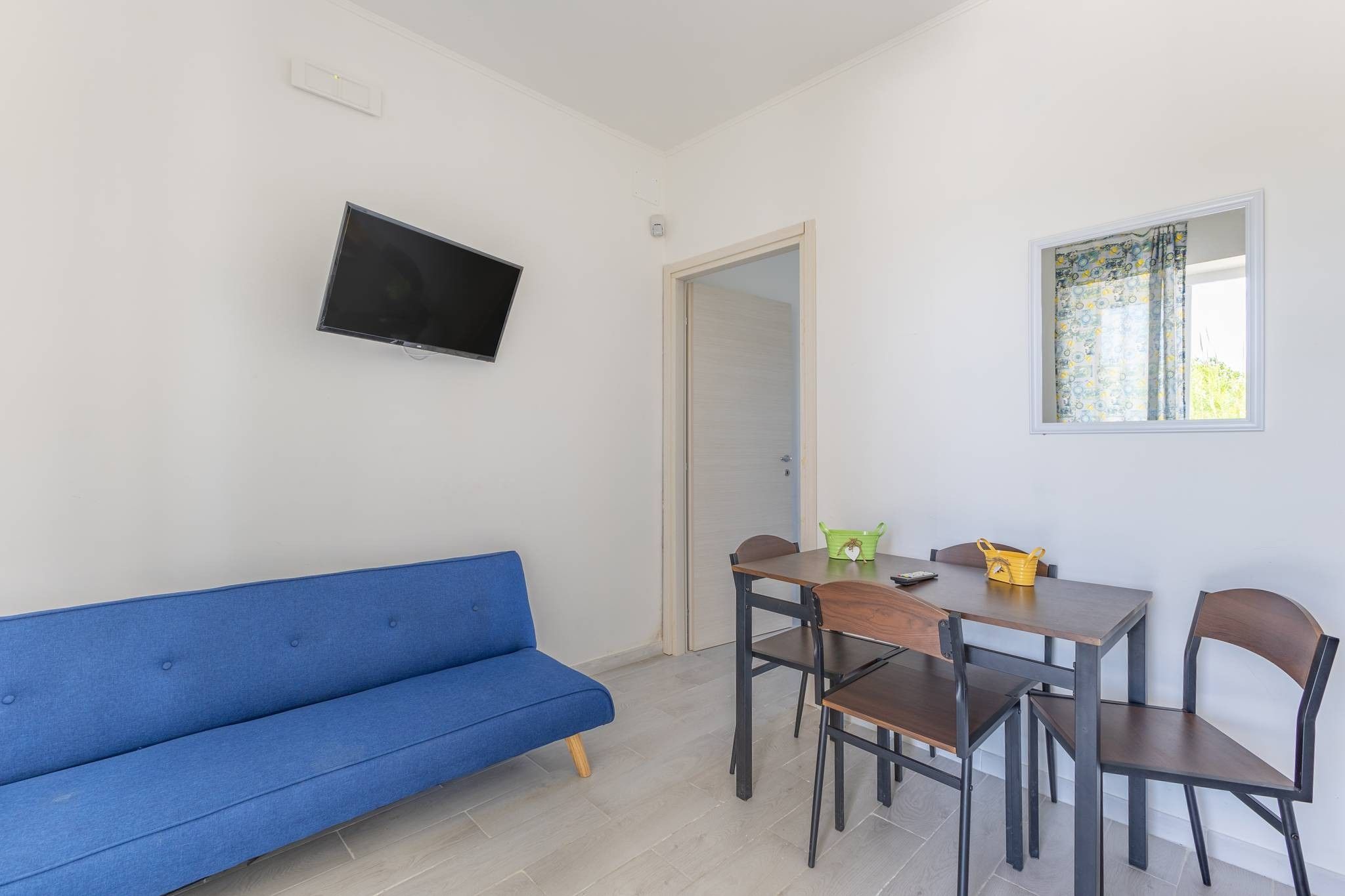 Una stanza con un divano blu, un tavolo da pranzo con sedie, una televisione a parete e una finestra con tende.