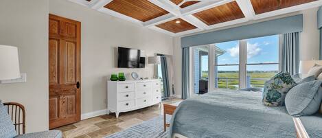 A bedroom with a large bed, a wooden ceiling, a white dresser, and a view of the outdoors.