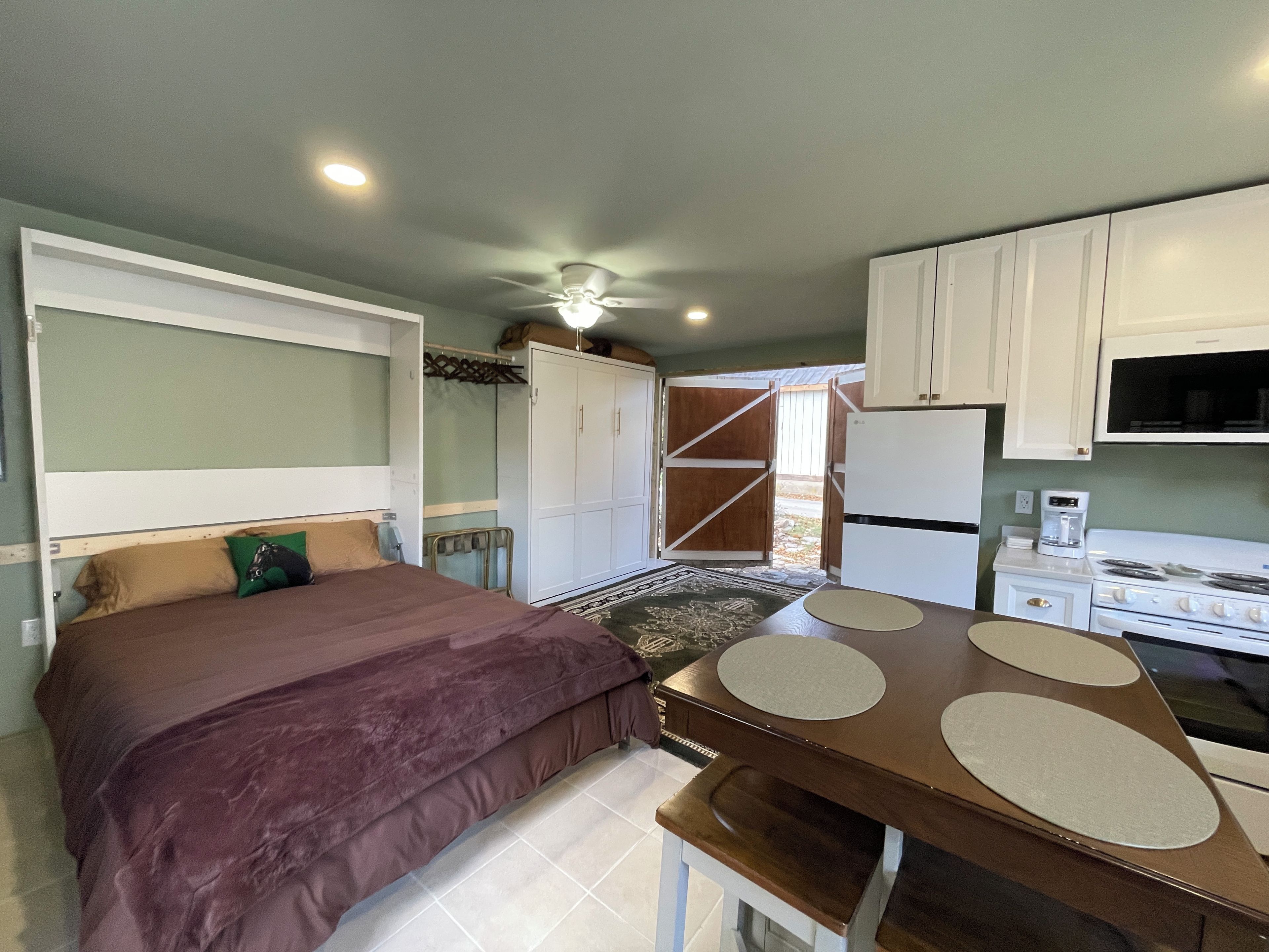 A compact bedroom with a single bed, a small table, and a kitchenette area with a stove and refrigerator.