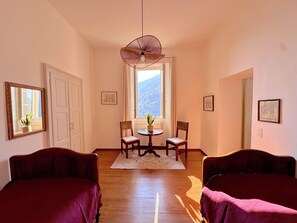 A hotel room with a wooden floor, two beds, a dining table with chairs, a hanging light fixture, and a window with a mountain view.