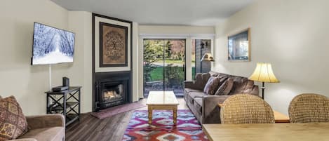 A living room with a fireplace, a television, a sofa, a coffee table, and a view through a glass door.