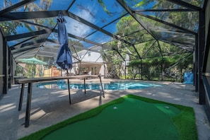 Zona de piscina con césped artificial verde para putting, mesa de billar y área de estar cubierta con sombrillas.
