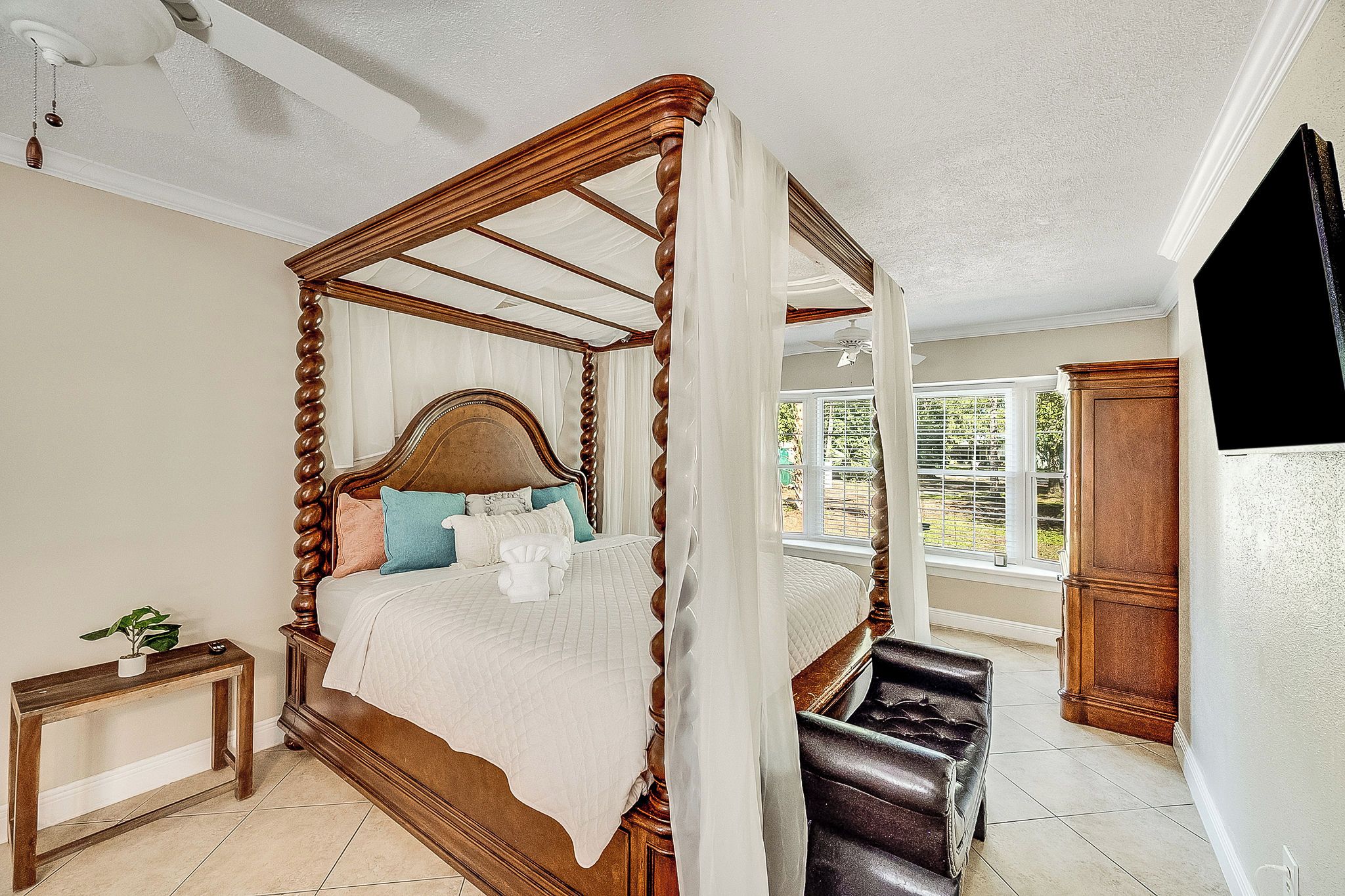 A four-poster bed with a canopy, a bedside table with a plant, and a view of the outdoors through a window.