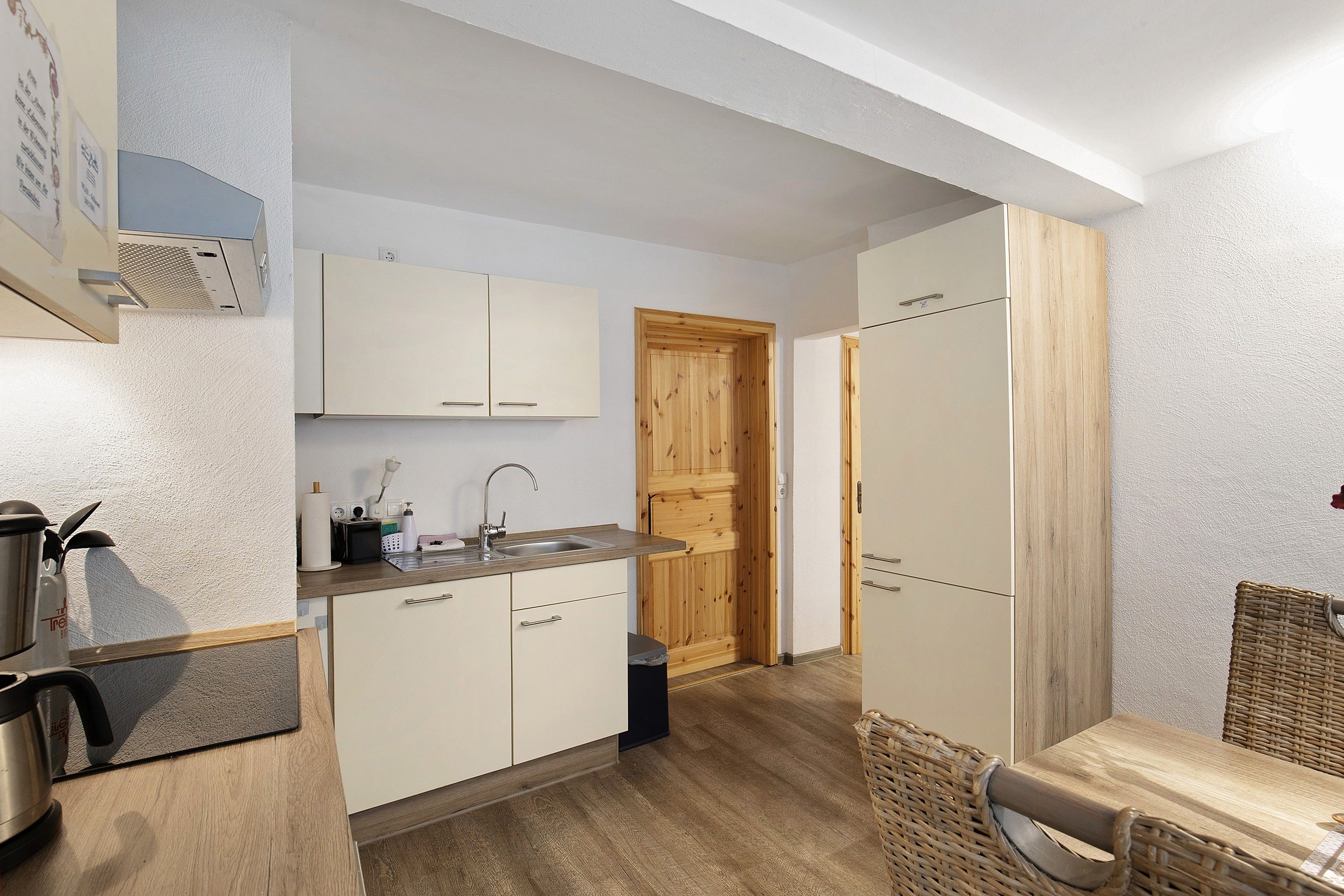 Una cucina compatta con mobili bianchi, una porta in legno e un piccolo angolo ristoro con un tavolino in vimini.