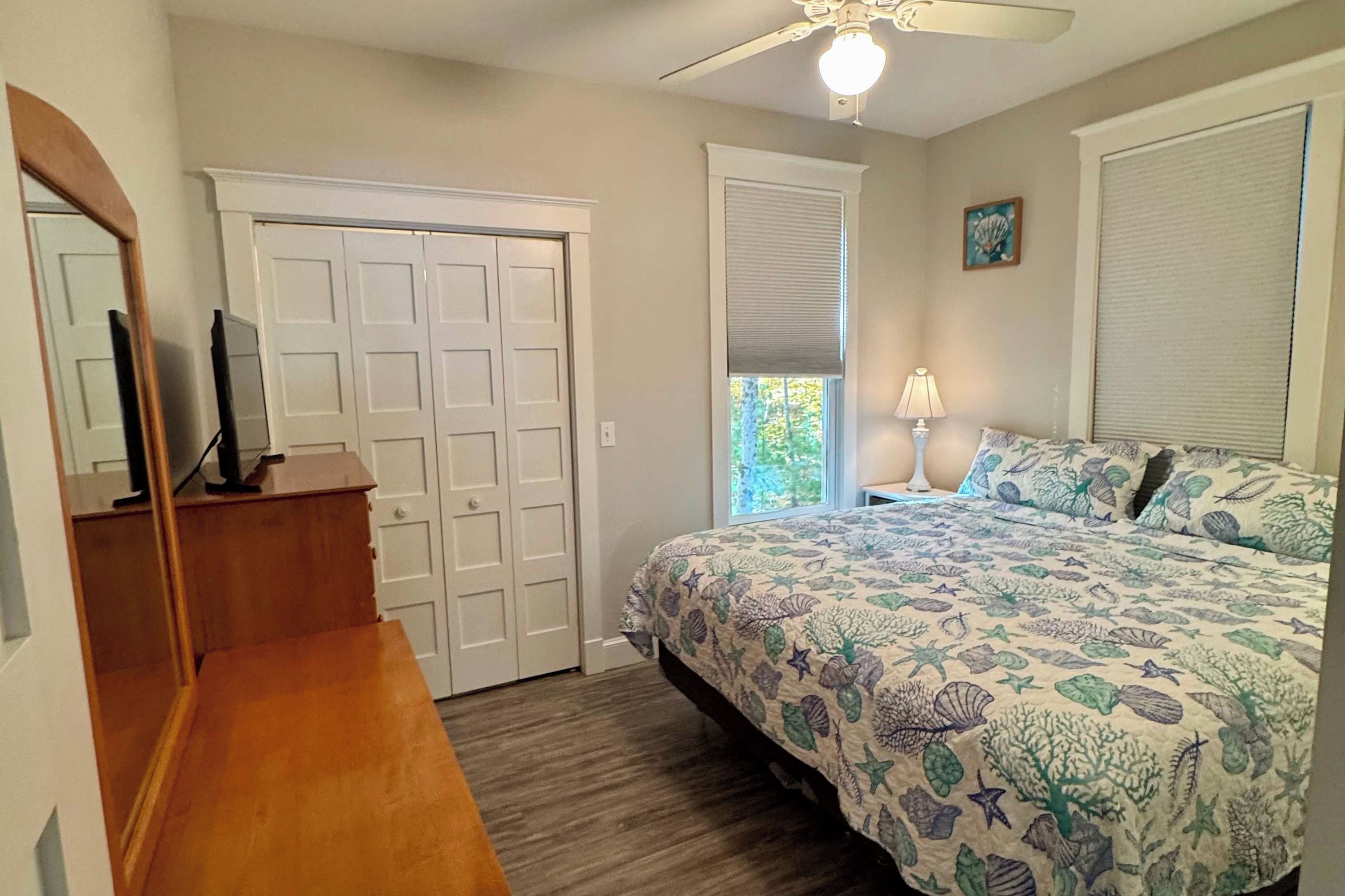 A bedroom with a bed, a dresser, a ceiling fan, a window with blinds, and a door.
