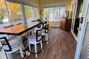 Un comedor con una mesa de madera y sillas blancas, un televisor sobre un mueble de madera y ventanas amplias con vistas a árboles y una calle.