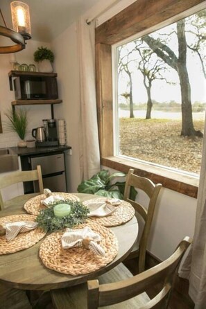 A dining area with a round table set for four, surrounded by wooden chairs. A window with a view of trees is visible. - Plush Cabin 4 min to Homestead Heritage 18 min to (Waco)