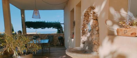 An outdoor patio area with a stone wall, a hanging light fixture, a table with chairs, and a view of the sea.