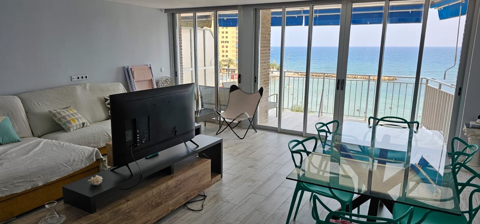 Een woonkamer met een flatscreen televisie op een houten standaard, een bank met kussens, een glazen eettafel met stoelen en een balkon met uitzicht op zee.