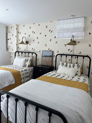 Quarto com duas camas, cada uma com cabeceira e mesa de cabeceira. Há um ar condicionado fixado na parede e um quadro na parede.