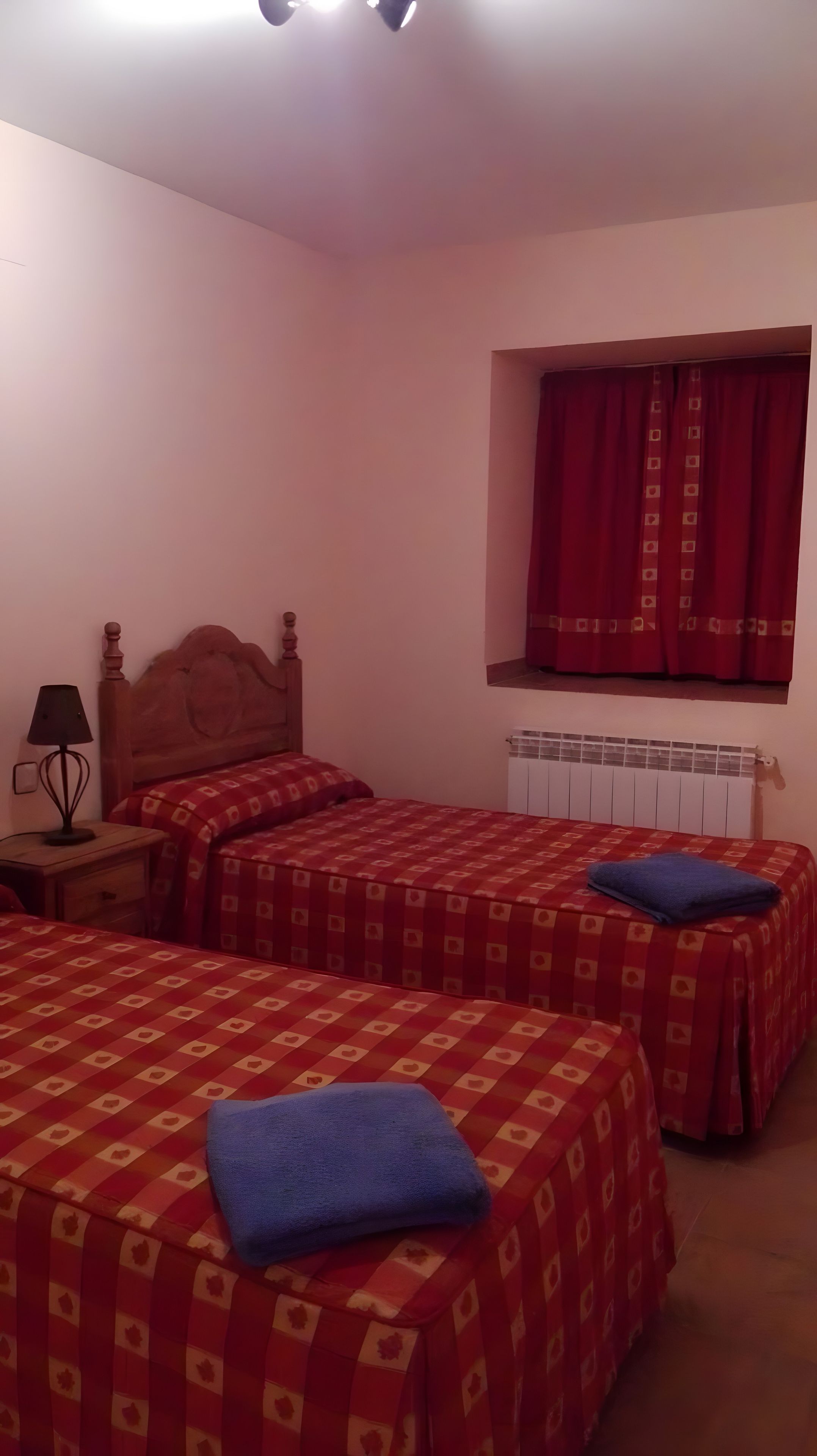Une chambre avec deux lits, une table de chevet, une lampe, une fenêtre avec des rideaux et un radiateur.